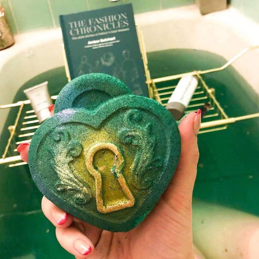 The Green bath bomb with a labradorite tumbler inside in front with it being used in a bath in a background